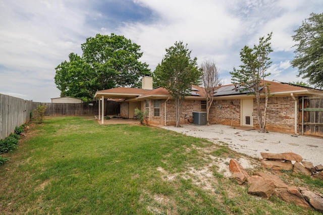 rear view of house with a fenced backyard, a chimney, central AC unit, roof mounted solar panels, and a lawn