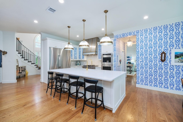 kitchen featuring wall chimney range hood, visible vents, appliances with stainless steel finishes, a center island with sink, and light countertops