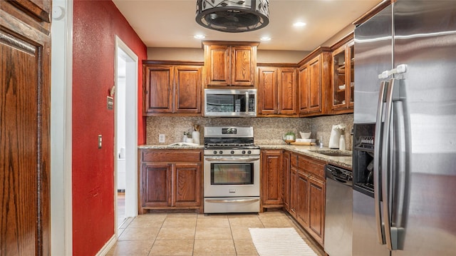 kitchen featuring brown cabinets, tasteful backsplash, light stone counters, light tile patterned floors, and stainless steel appliances
