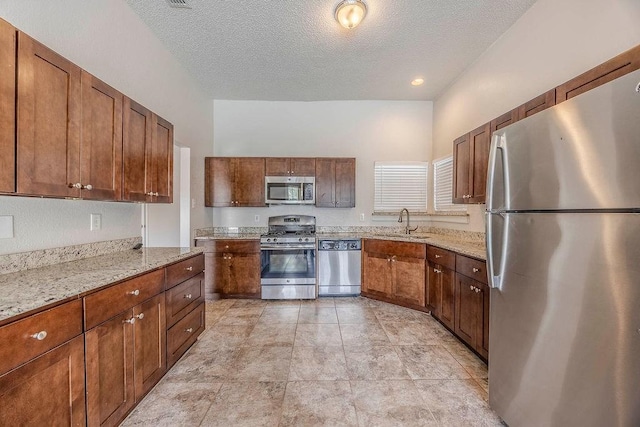 kitchen with brown cabinetry, a textured ceiling, light stone counters, a sink, and stainless steel appliances