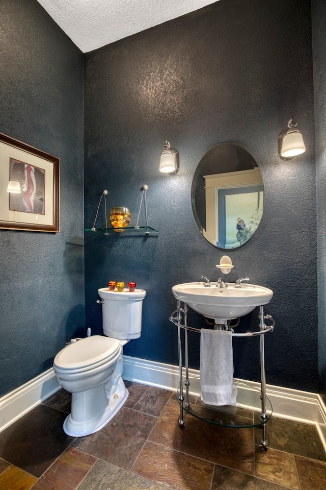half bathroom with stone tile floors, toilet, baseboards, and a textured ceiling