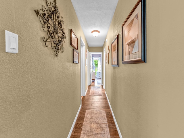corridor featuring a textured ceiling, a textured wall, baseboards, and dark wood-type flooring