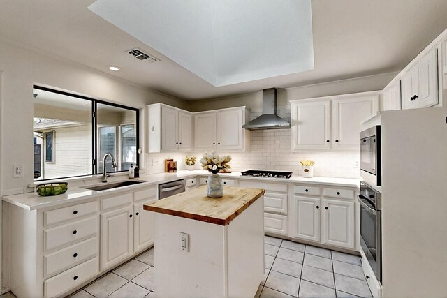 kitchen with wall chimney exhaust hood, butcher block counters, a sink, appliances with stainless steel finishes, and white cabinetry