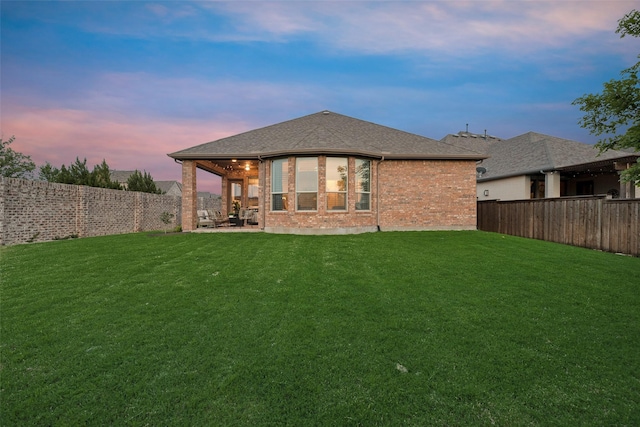 back of property at dusk with a lawn, brick siding, a fenced backyard, a patio, and a shingled roof