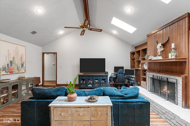 living room featuring vaulted ceiling with skylight, a fireplace, a ceiling fan, light wood-type flooring, and visible vents