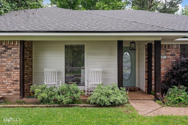 view of exterior entry with brick siding, roof with shingles, and covered porch