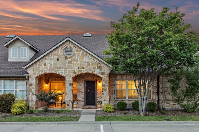 french country inspired facade featuring roof with shingles, covered porch, and stone siding