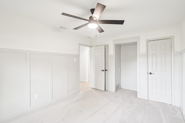unfurnished bedroom featuring a ceiling fan, light colored carpet, visible vents, wainscoting, and a decorative wall