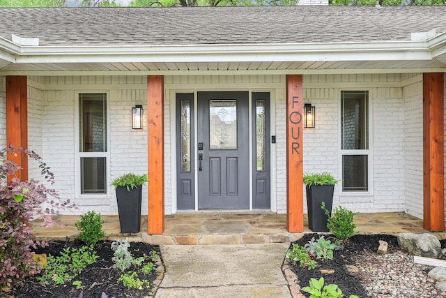view of exterior entry with a shingled roof, a porch, and brick siding