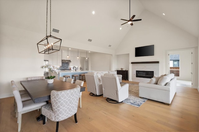 living room featuring visible vents, light wood-style floors, high vaulted ceiling, ceiling fan with notable chandelier, and a premium fireplace