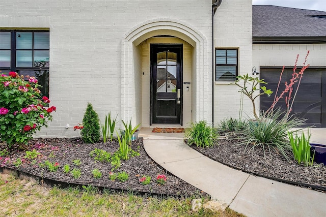 view of exterior entry featuring a shingled roof, an attached garage, and brick siding