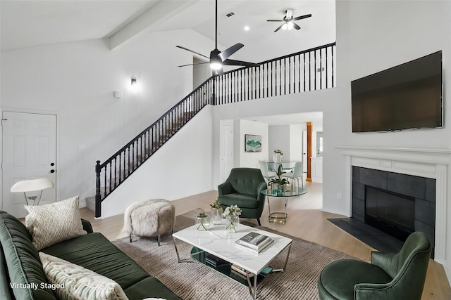 living area featuring a ceiling fan, a tiled fireplace, wood finished floors, high vaulted ceiling, and stairs