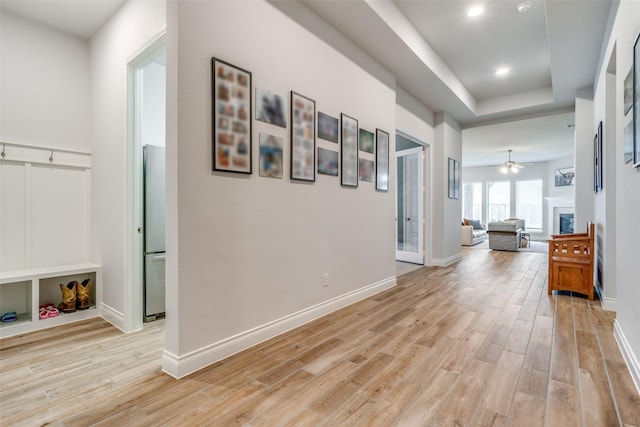 corridor featuring light wood-style floors, recessed lighting, baseboards, and a raised ceiling