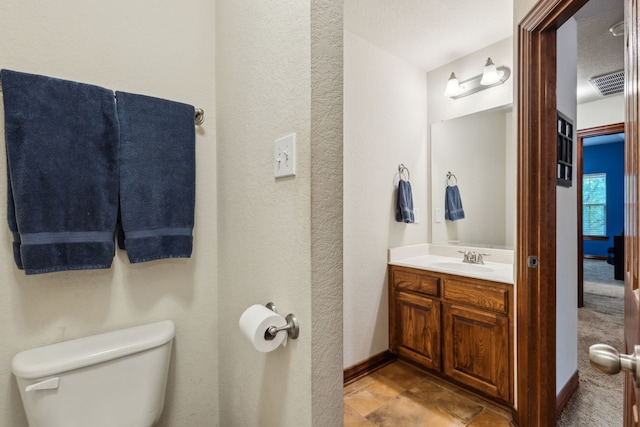 half bath featuring vanity, visible vents, a textured wall, toilet, and baseboards