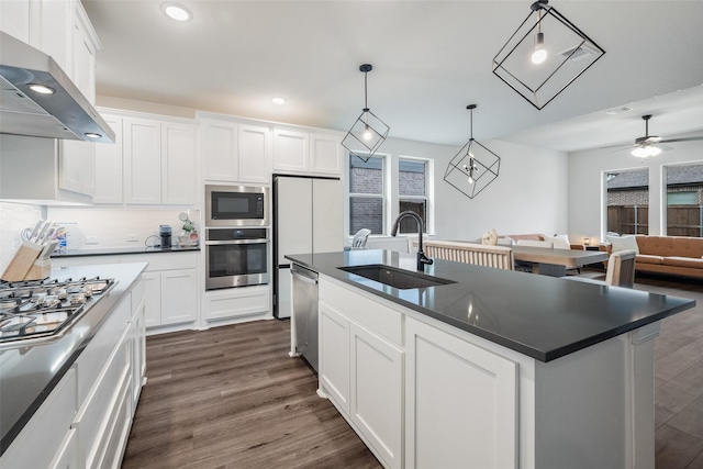 kitchen featuring appliances with stainless steel finishes, dark countertops, a sink, recessed lighting, and wall chimney exhaust hood
