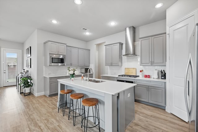 kitchen with a center island with sink, wall chimney exhaust hood, a sink, gray cabinetry, and stainless steel appliances