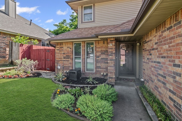 view of exterior entry with a yard, brick siding, fence, and roof with shingles