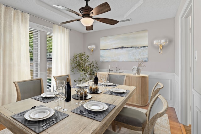 dining area with a wainscoted wall, a ceiling fan, light wood finished floors, and visible vents