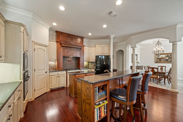 1028 Gabriel Ln, Fort Worth, TX 76116 - kitchen featuring a kitchen island with sink, black appliances, decorative columns, cream cabinets, and dark wood-style flooring