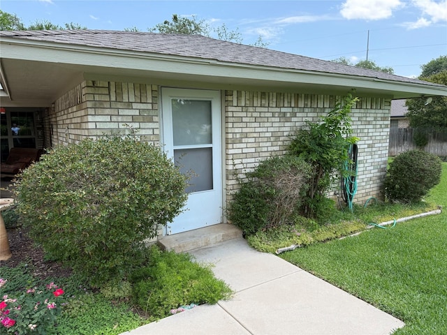 view of exterior entry featuring brick siding, a shingled roof, and a yard