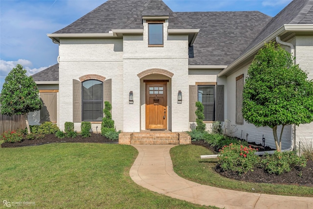 french country inspired facade featuring brick siding, a shingled roof, and a front yard