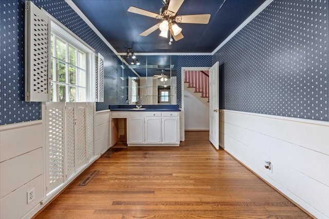 unfurnished dining area featuring ornamental molding, wallpapered walls, a wainscoted wall, a ceiling fan, and light wood finished floors