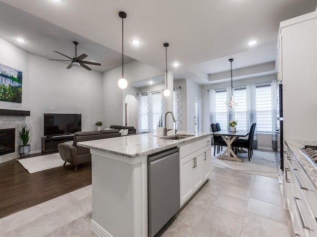kitchen with a ceiling fan, a tiled fireplace, stainless steel dishwasher, a sink, and light stone counters