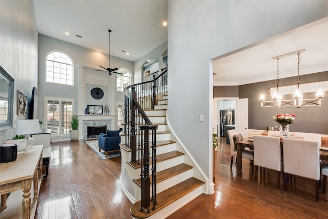staircase featuring wood finished floors, baseboards, a high ceiling, a tile fireplace, and ceiling fan with notable chandelier
