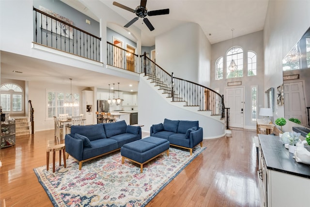 living room featuring stairs, light wood-type flooring, ceiling fan with notable chandelier, and a towering ceiling