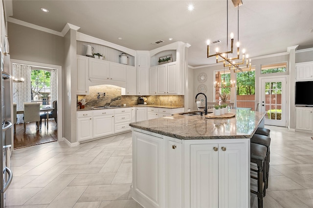kitchen featuring an inviting chandelier, decorative backsplash, a kitchen island with sink, dark stone counters, and a sink