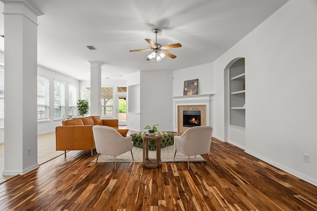 living room with built in shelves, a tiled fireplace, a ceiling fan, dark wood-type flooring, and ornate columns