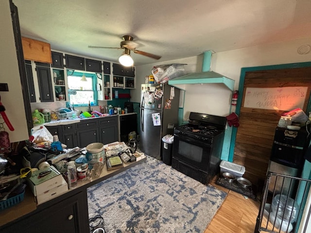 kitchen with black gas range, freestanding refrigerator, ceiling fan, wall chimney range hood, and light wood finished floors