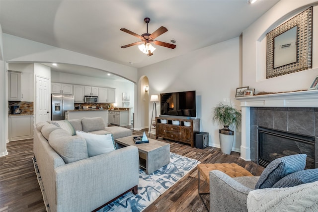 living room featuring baseboards, ceiling fan, a tiled fireplace, arched walkways, and dark wood finished floors