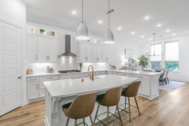 kitchen with wall chimney range hood, a peninsula, an island with sink, and light countertops