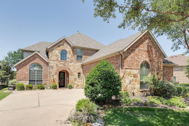 french country style house featuring roof with shingles, driveway, brick siding, and stone siding