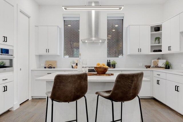 kitchen featuring open shelves, oven, light wood-style floors, a breakfast bar area, and wall chimney range hood