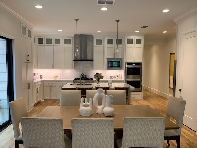 kitchen featuring ornamental molding, a kitchen island with sink, wall chimney exhaust hood, light wood finished floors, and appliances with stainless steel finishes