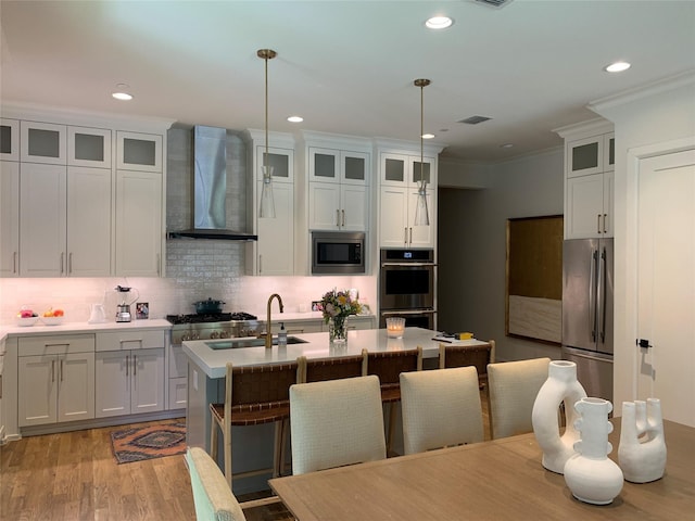 kitchen with wall chimney range hood, an island with sink, stainless steel appliances, decorative backsplash, and wood finished floors