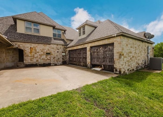 back of house with a shingled roof, stone siding, central AC unit, a yard, and driveway
