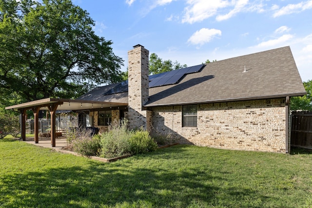 back of house with a chimney, a lawn, fence, roof mounted solar panels, and a shingled roof