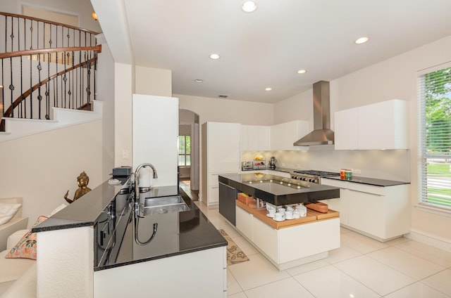 kitchen featuring white cabinets, dark countertops, a sink, plenty of natural light, and wall chimney exhaust hood