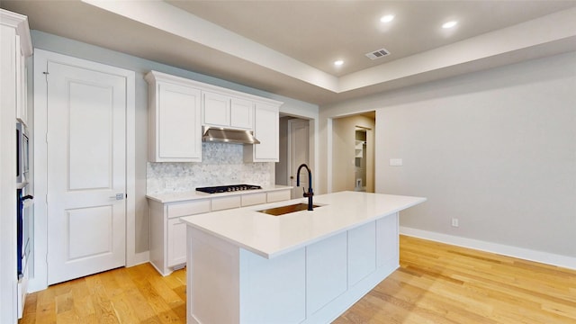 kitchen with a sink, light countertops, decorative backsplash, light wood-type flooring, and under cabinet range hood