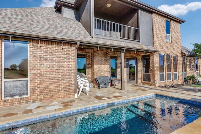 rear view of property with roof with shingles, brick siding, an outdoor pool, a balcony, and a patio