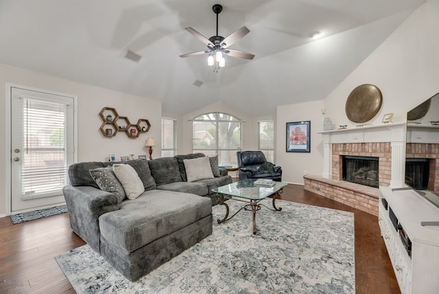 living room featuring a ceiling fan, visible vents, vaulted ceiling, a brick fireplace, and dark wood finished floors