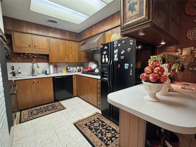 kitchen featuring brown cabinets, a sink, light countertops, wall chimney exhaust hood, and black dishwasher