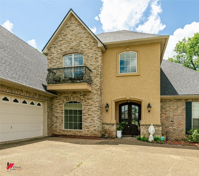 view of front facade with a balcony, stone siding, french doors, a shingled roof, and concrete driveway