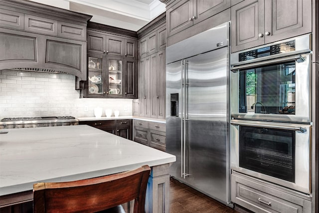 kitchen featuring light stone countertops, dark wood finished floors, stainless steel appliances, backsplash, and glass insert cabinets