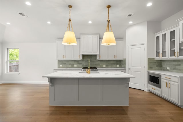 kitchen with an island with sink, stainless steel microwave, decorative backsplash, visible vents, and pendant lighting