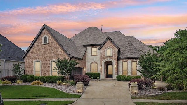 french country home with roof with shingles, concrete driveway, a yard, and brick siding