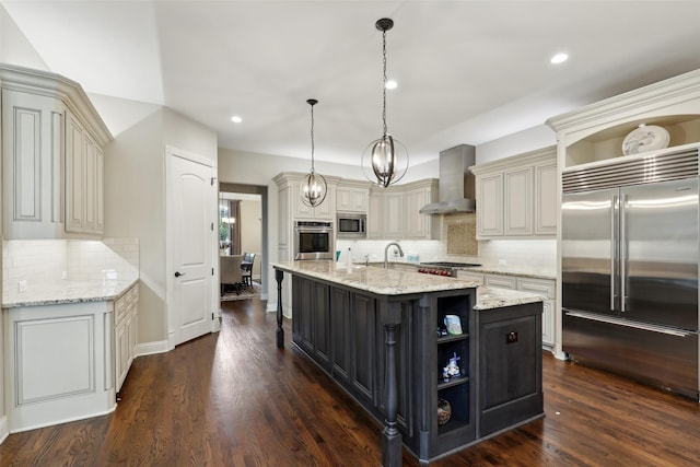 kitchen with an island with sink, open shelves, wall chimney exhaust hood, and built in appliances